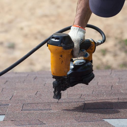 Roofer With A Nail Gun Securing Asphalt Shingles on a New House Under Construction